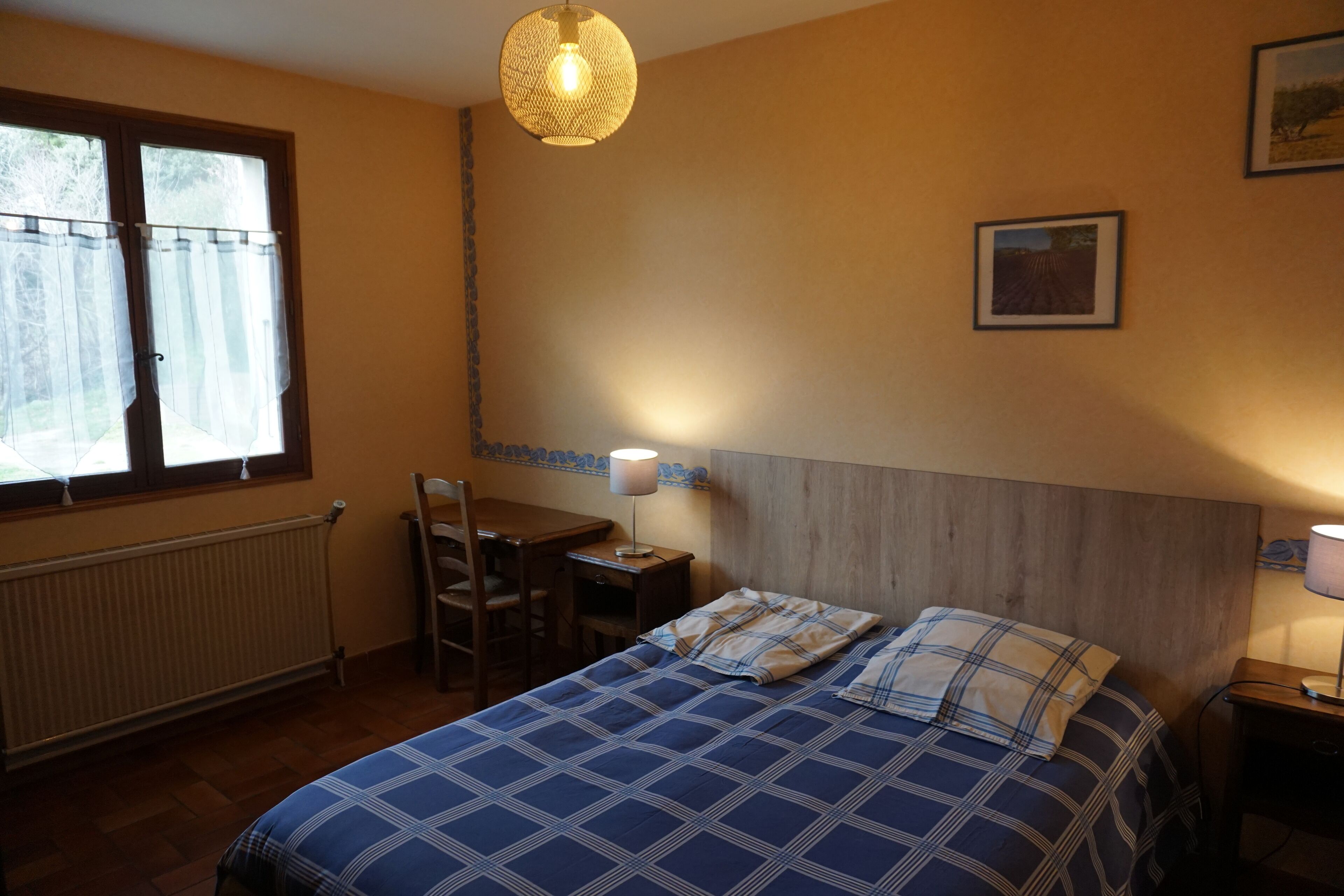 Een slaapkamer met een bed, een bureau, een stoel, een lamp en een raam met gordijnen.