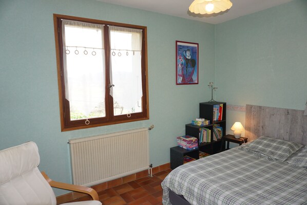 Une chambre à coucher comprenant un lit, une chaise, un radiateur, une fenêtre avec des rideaux, une bibliothèque et un tableau accroché au mur.