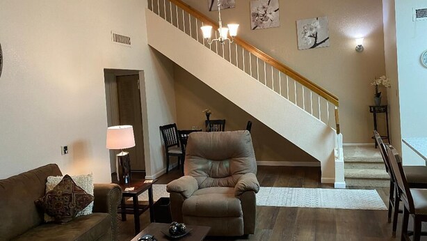 A living room with a beige sofa, a recliner, a coffee table, and a lamp. There is a staircase with a chandelier and artwork on the walls. - Your Perfect Getaway Awaits- Ideal for Students and Families (Chandler)