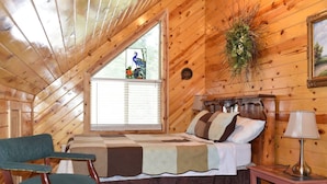Um quarto aconchegante com teto inclinado de madeira, cama com roupa de cama estampada, uma cadeira, mesa de cabeceira com abajur e janela com persianas.