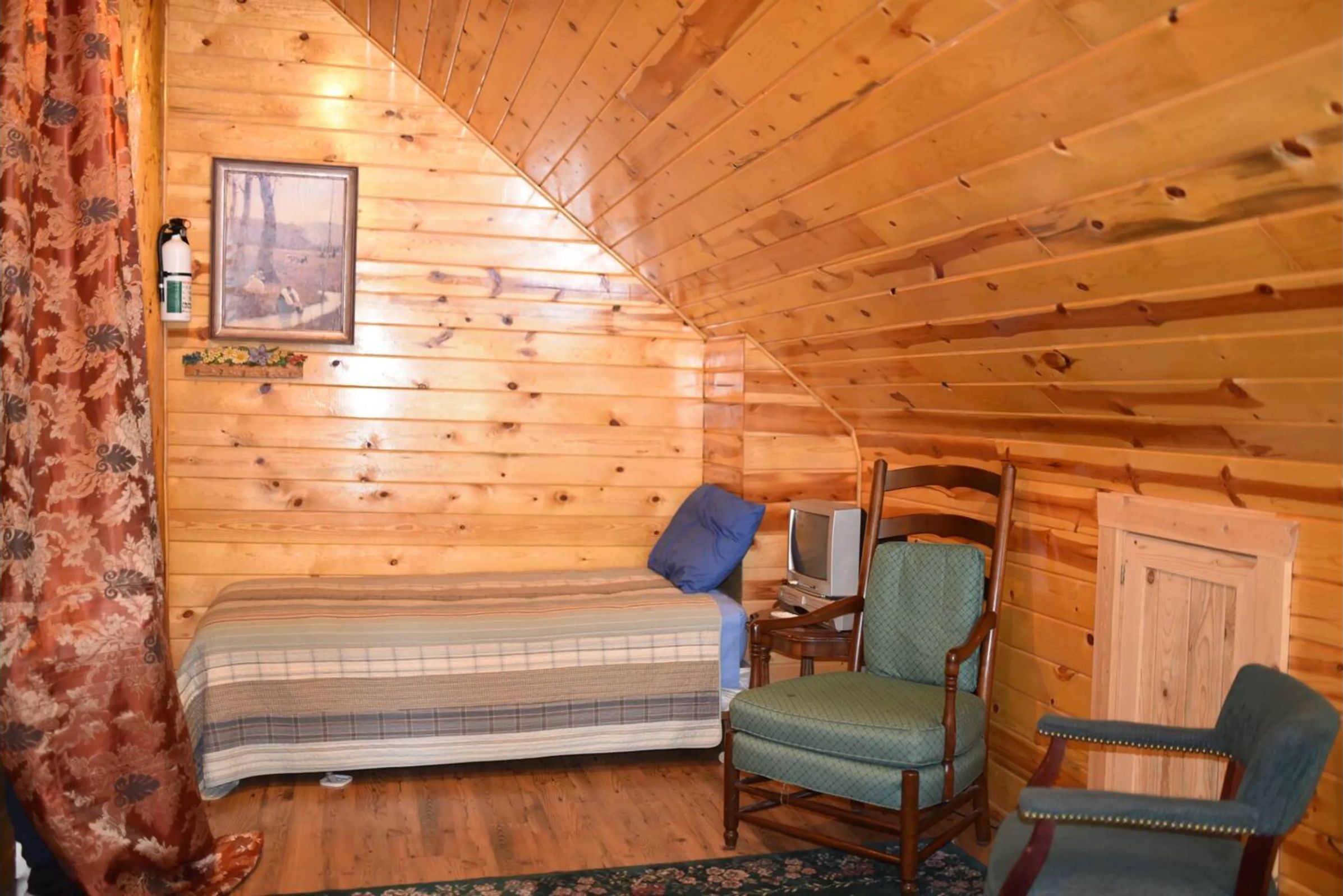 Una habitación de cabaña acogedora, con cama, sillón, televisor y un cuadro en la pared.