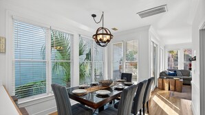 Uma sala de jantar com uma mesa grande posta para seis pessoas, rodeada por cadeiras, e vista para o exterior através de amplas janelas.