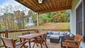A wooden deck with a table and chairs, a sofa, and a window overlooking a yard with trees and a fence.