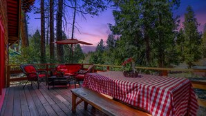 A wooden deck with a checkered table and red chairs, surrounded by tall trees and a sunset view. - 5 STAR **STRAWBERRY CORNER** CLASSIC CABIN ON CREEK, 1 MINUTE WALK TO VILLAGE! (Idyllwild)