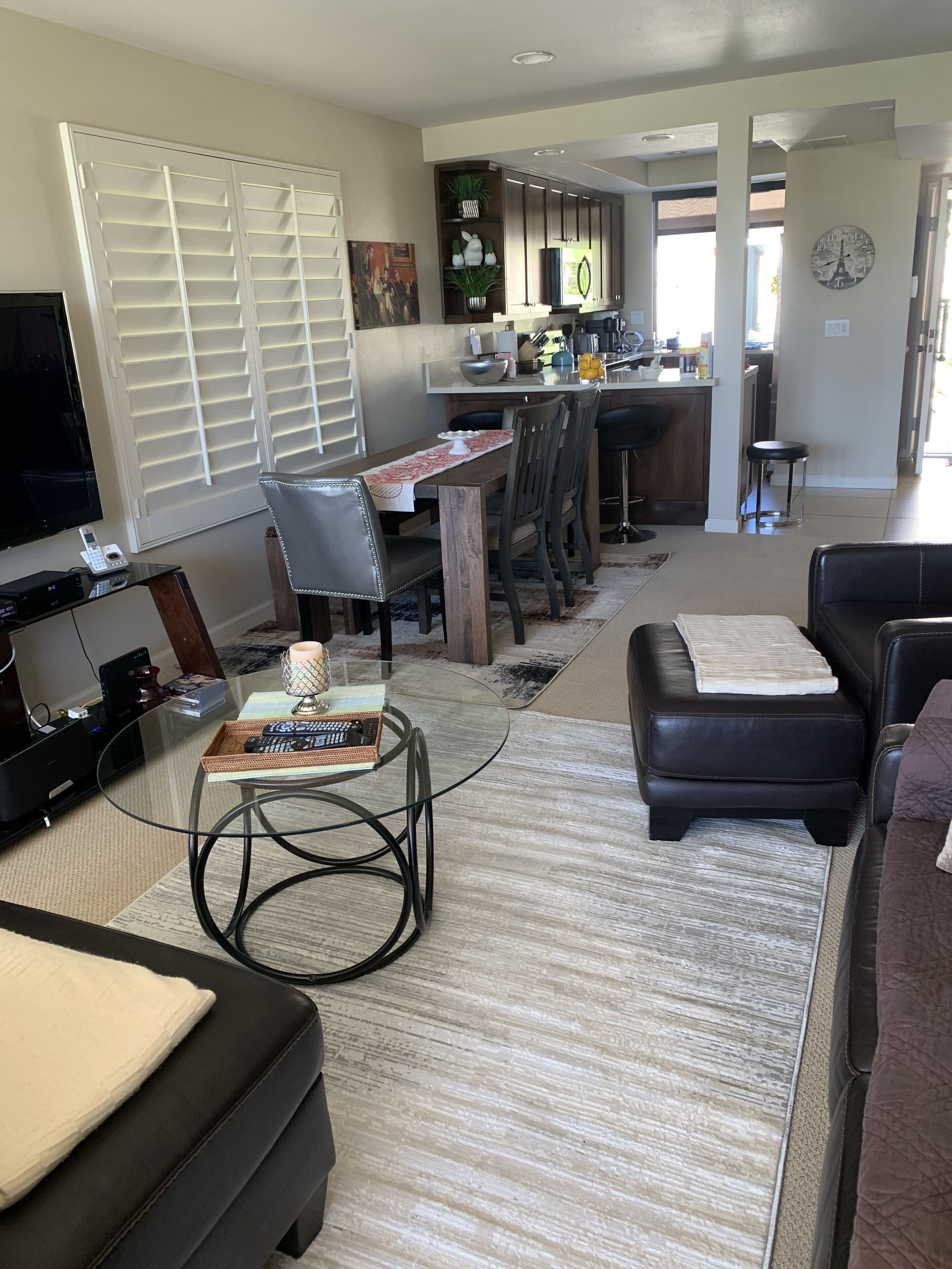 Uma sala de estar moderna com televisão, sofá e mesa de centro de vidro. Há uma área de refeições com cadeiras e uma cozinha ao fundo.
