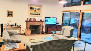 A cozy living room with a fireplace, comfortable seating, a television, and a large window offering a view of the outdoors.
