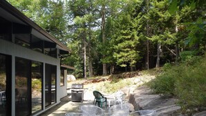 An outdoor patio with metal chairs and a grill, surrounded by a forested area.