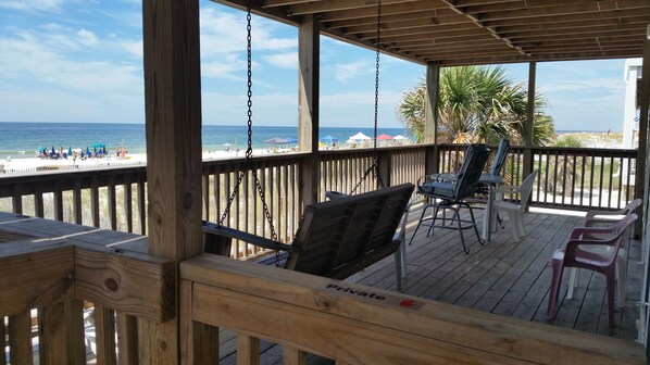 A wooden deck with a swing chair and a table, overlooking a beach with umbrellas and people.
