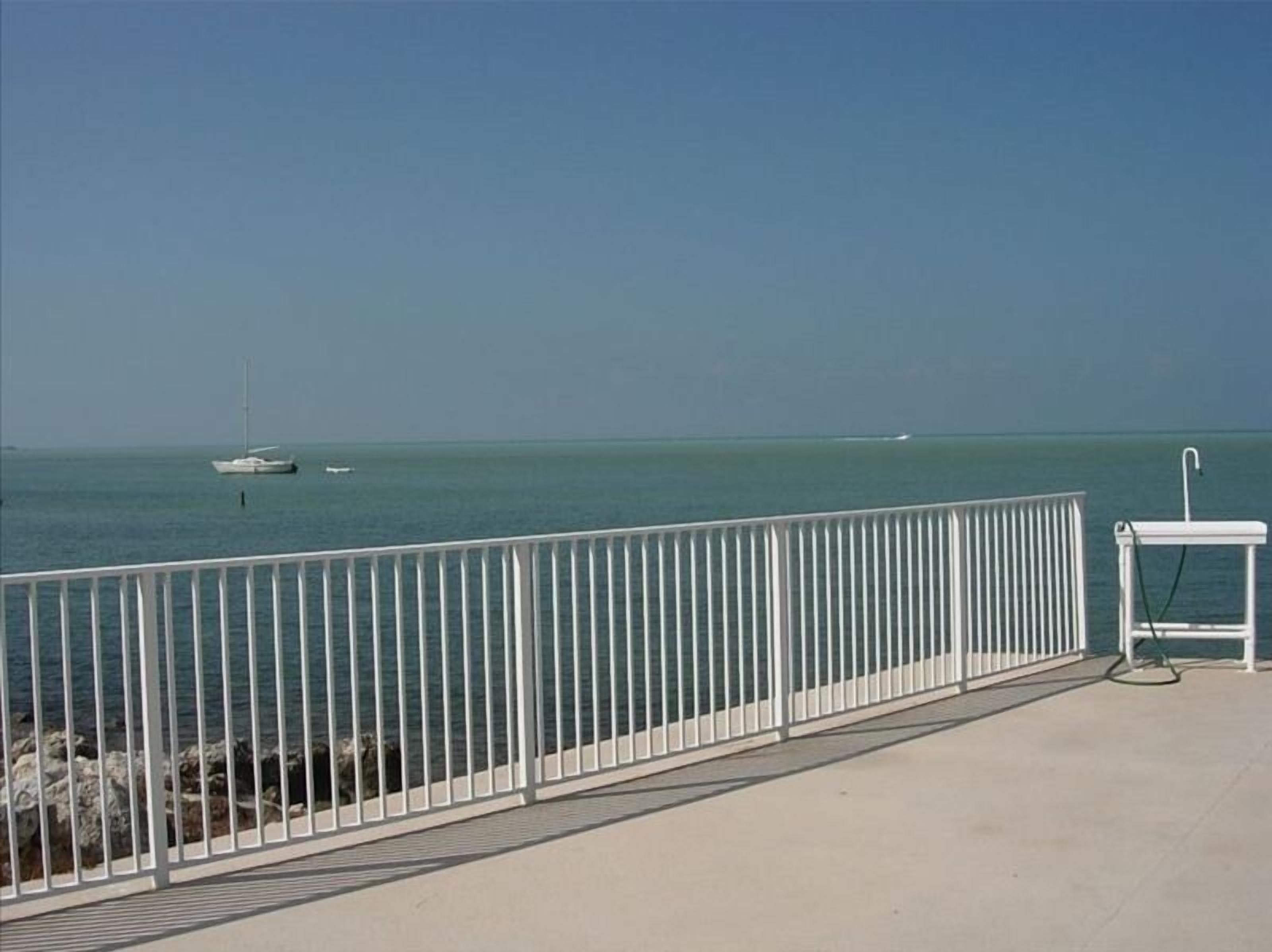 A coastal walkway with a white railing, a small dock with a sailboat, and a white structure with a hose.