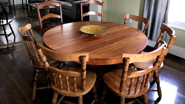 Una mesa de comedor redonda de madera con seis sillas, un plato con borde dorado y un sofá verde al fondo.
