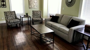 Una sala de estar con un sofá, dos sillones, una mesita de centro y una mesita auxiliar. Hay dos ventanas con persianas, un reloj de pared y un espejo decorativo.