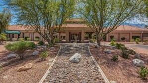 A building with a tiled roof and a covered entrance. There are trees and a gravel pathway leading to the entrance. - Desert Princess Cc And Resort, On Golf Course, W/ Mountain Views (Cathedral City)