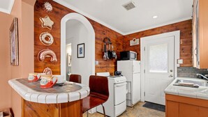 Una cocina con paredes de madera, un refrigerador blanco, una estufa y un microondas. Hay una silla roja y una mesa con encimera blanca.
