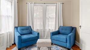 Una habitación con dos sillones azules, un otoman blanco y un ventilador de techo.