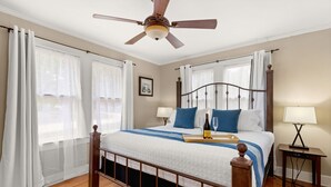 Un dormitorio con cama, dos mesitas de noche, ventilador de techo y ventana con cortinas.