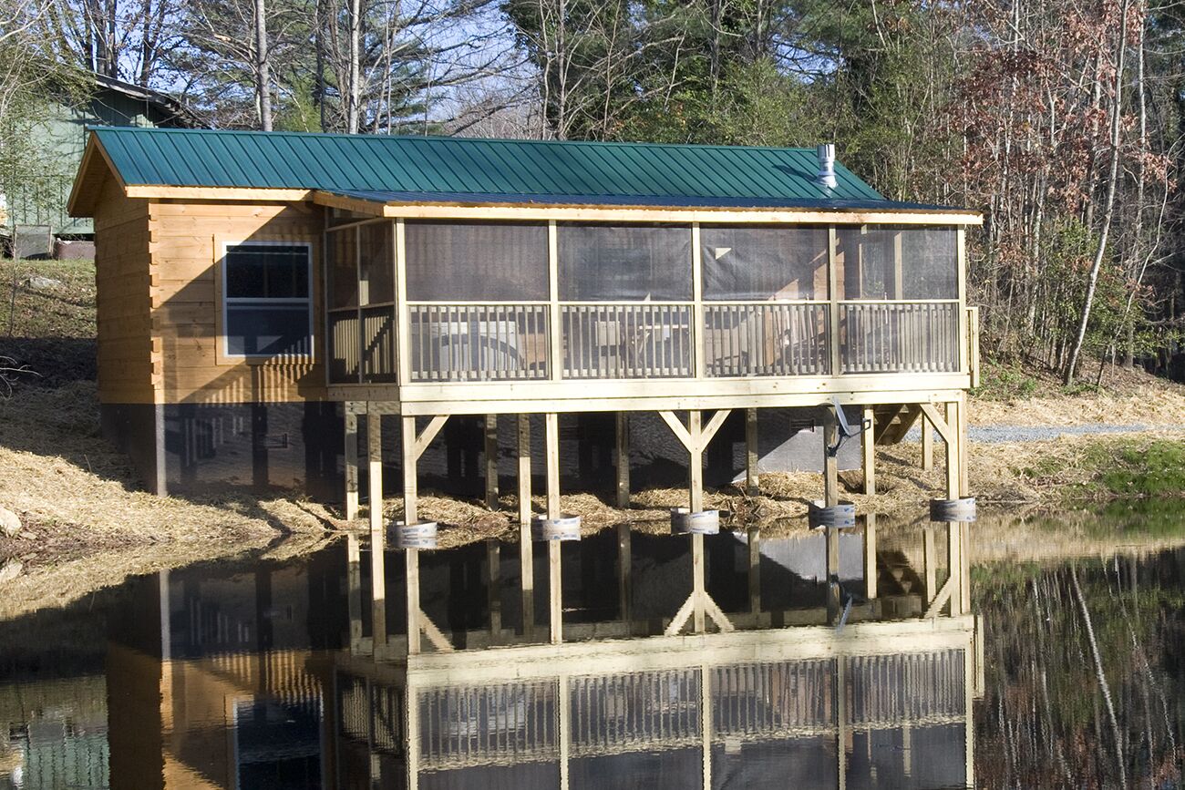 A wooden cabin with a green roof, a balcony, and a dock extending into a calm body of water.