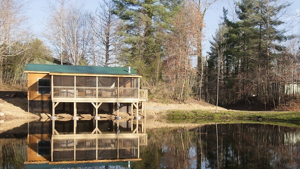 Une cabane en bois avec un toit vert, une terrasse et son reflet dans une étendue d’eau calme.