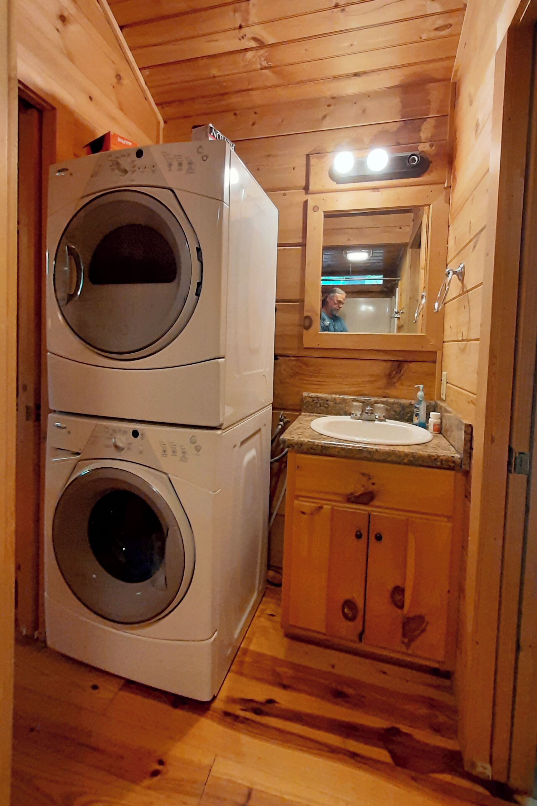 Une petite buanderie équipée de deux machines à laver superposées, d’un évier et de meubles de rangement en bois.