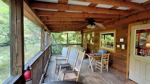 Une terrasse en bois avec des sièges d’extérieur, un ventilateur de plafond et une vue sur la forêt environnante.