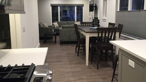 Eine moderne Küche mit Essbereich, ein Wohnzimmer mit Sofa und Fernseher sowie große Fenster mit Jalousien.