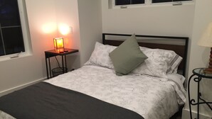 Ein ordentlich bezogenes Bett mit floralem Bettzeug, ein Nachttisch mit Lampe und ein Fenster mit Blick auf Bäume.