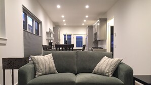 Ein modernes Wohnzimmer mit einem grauen Sofa, einem Glastisch und einem Essbereich im Hintergrund.
