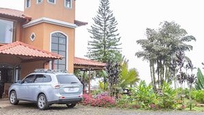 Un bâtiment avec un toit recouvert de tuiles rouges, une voiture garée devant et diverses plantes tropicales.