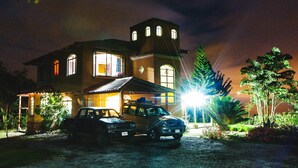 Une grande maison avec une tour, deux véhicules garés et un jardin bien éclairé.