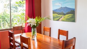 Un coin repas avec une table en bois, des chaises, un vase de fleurs, un tableau représentant un paysage et une vue sur la verdure par la fenêtre.
