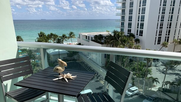 En balkon med et bord og stole, der ud til et pool og udsigt over havet og en bygning.