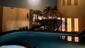 A nighttime view of a swimming pool area with a moon in the sky, palm trees, and a building with illuminated windows.