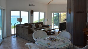 A living room with a sofa, two chairs, a coffee table, and a TV. There are large windows with a view of the ocean. - Oceanfront Glass House on  Deserted Pink Sand Beach-directly on beach!  (Governors Harbour)