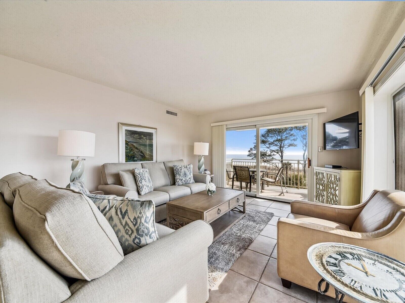 A living room with a sofa, coffee table, and a TV. There is a balcony with a view of the ocean.