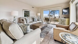 Ein Wohnzimmer mit Sofa, Couchtisch und Fernseher. Es gibt einen Balkon mit Blick auf den Ozean.