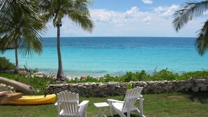 Zona verde con dos sillas Adirondack blancas mirando a una playa de aguas turquesas cristalinas y una palmera.