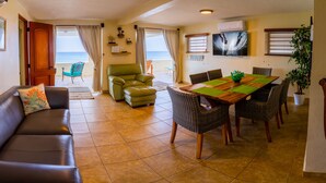 Un salón acogedor con un sofá de cuero marrón, un sillón verde y una mesa de comedor de madera con sillas de mimbre. Un ventanal ofrece vistas al océano.
