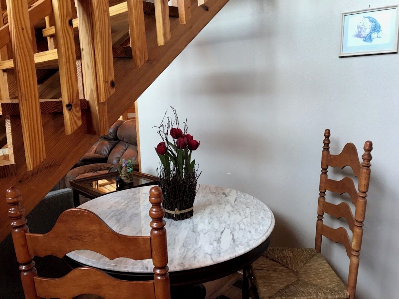 Un comedor con una mesa redonda de mármol, sillas de madera, un jarrón con flores rojas y una escalera al fondo.