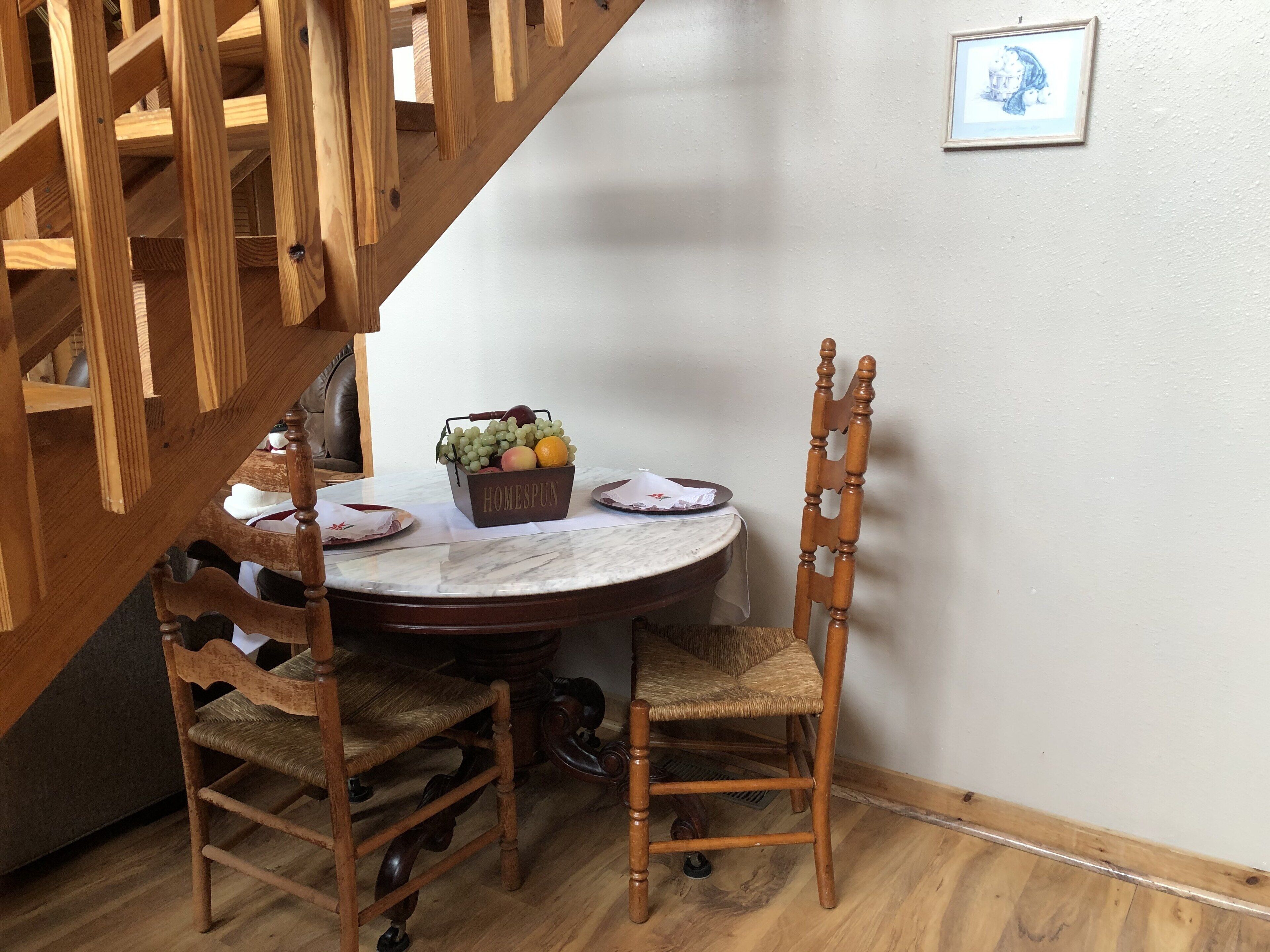 A dining area with a marble table, two wooden chairs, a fruit basket, and a staircase.
