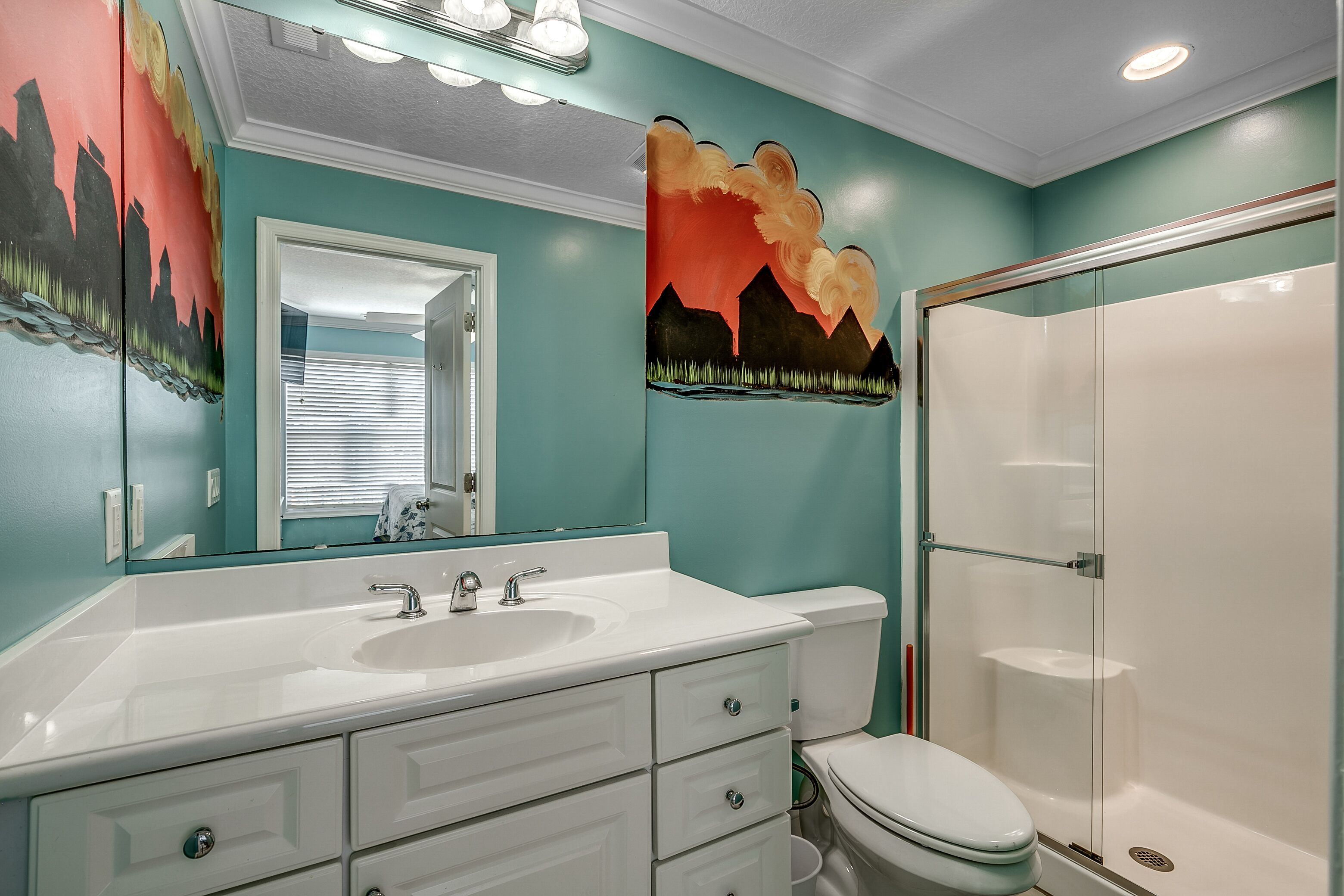 Un baño con lavamanos blanco, espejo y retrete, paredes color turquesa y zona de ducha.