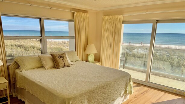 A bedroom with a large bed, a nightstand, a lamp, and a view of the beach through large windows.