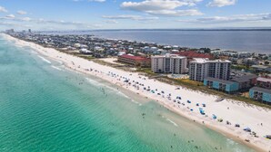 An aerial view of a coastal town with a long sandy beach, several beachfront buildings, and a clear view of the ocean.