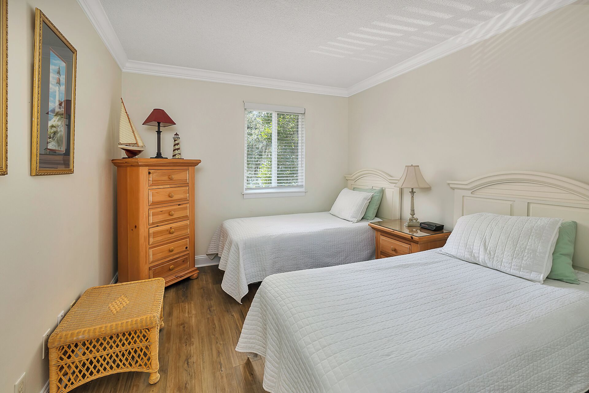 A bedroom with two single beds, a wooden dresser, a wicker chair, and a window with blinds.