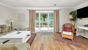 A living room with a white sofa, a wicker chair, a coffee table, and a view of a garden through a glass door. - YOUR ISLAND DESTINATION IS WAITING! (Saint Simons Island)