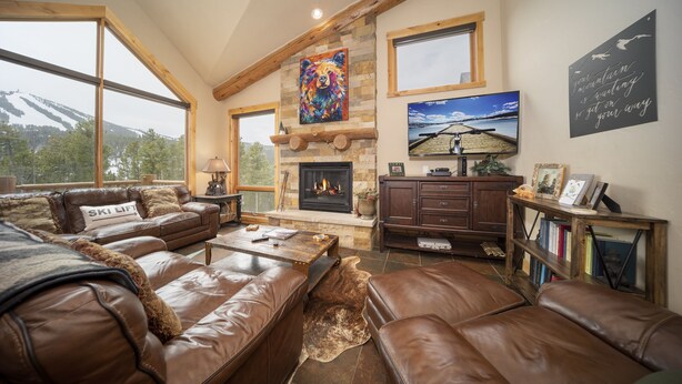 A cozy living room with leather sofas, a fireplace, a television, and a large window overlooking a snowy mountain.