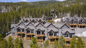 A row of modern log cabin-style buildings with balconies, surrounded by dense forest and a mountainous backdrop.