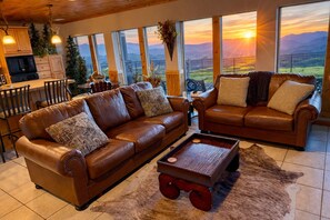 Un espacio de sala acogedor con sofás de cuero, una mesa de centro y vista a una puesta de sol sobre montañas.