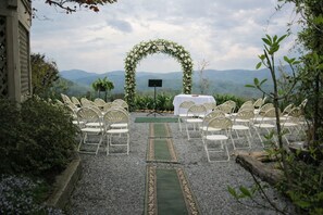 Un espacio para bodas al aire libre con un arco floral, asientos dispuestos y una mesa con una pantalla.