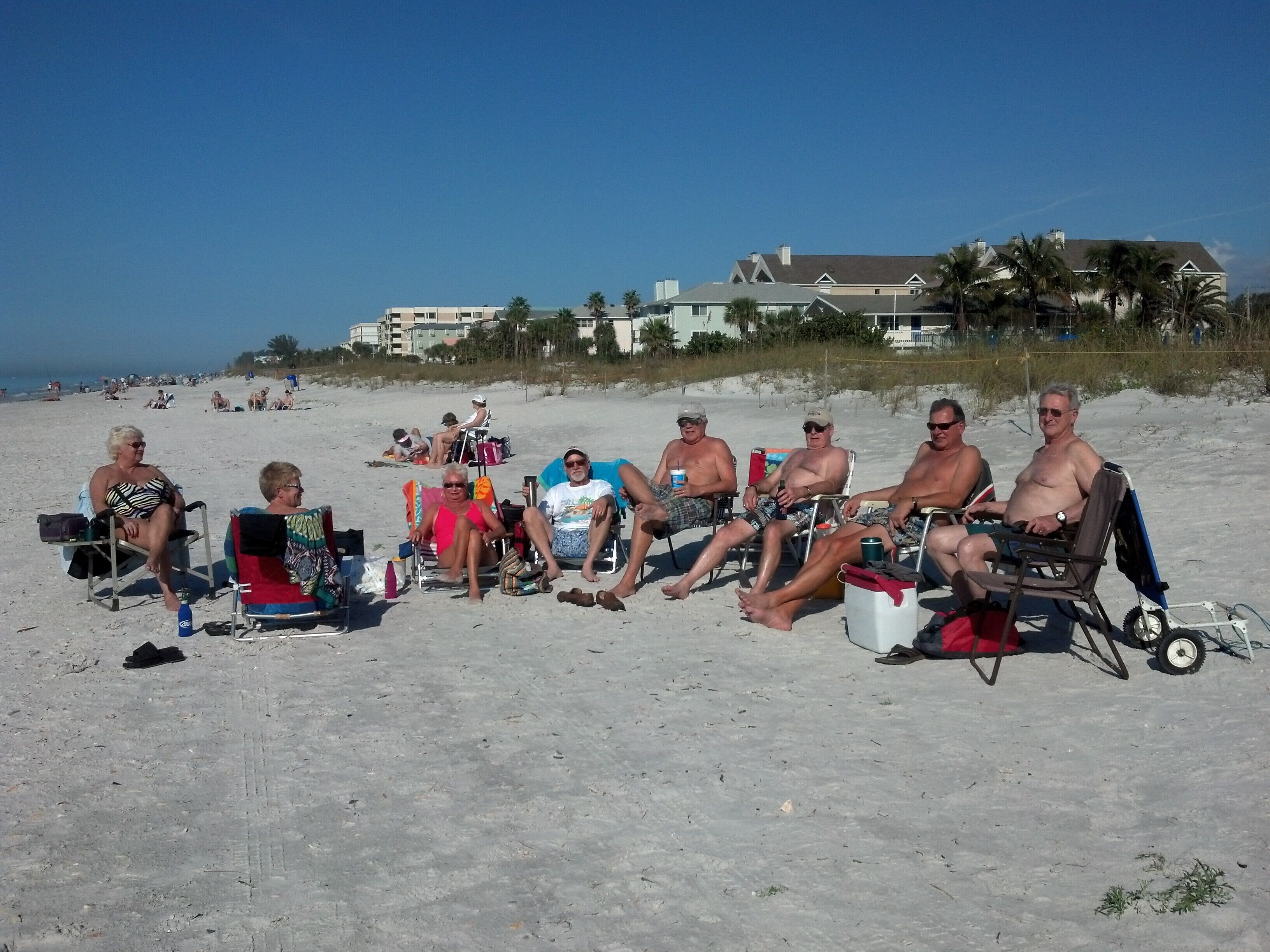 Um grupo de pessoas sentadas em cadeiras de praia em uma areia, com edifícios ao fundo.