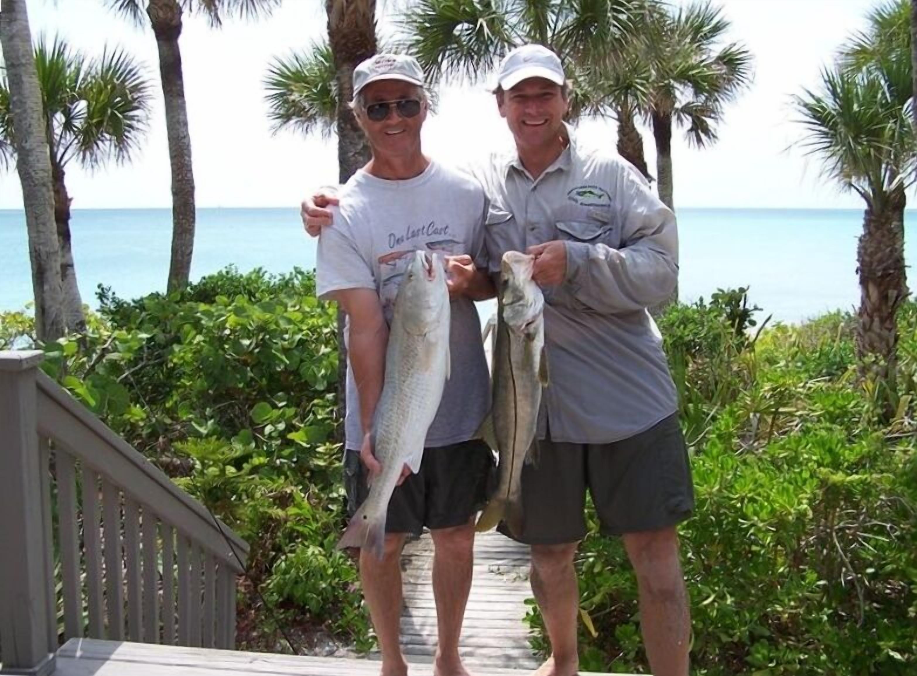 Two individuals holding fish on a wooden walkway with palm trees and a view of the ocean.