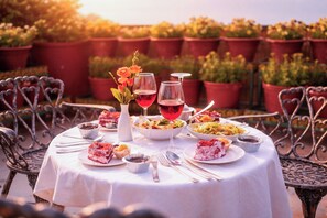 Mesa de comedor al aire libre con mantel blanco, rodeada de sillas metálicas, dispuesta con platos de comida, copas de vino tinto y un florero con flores.